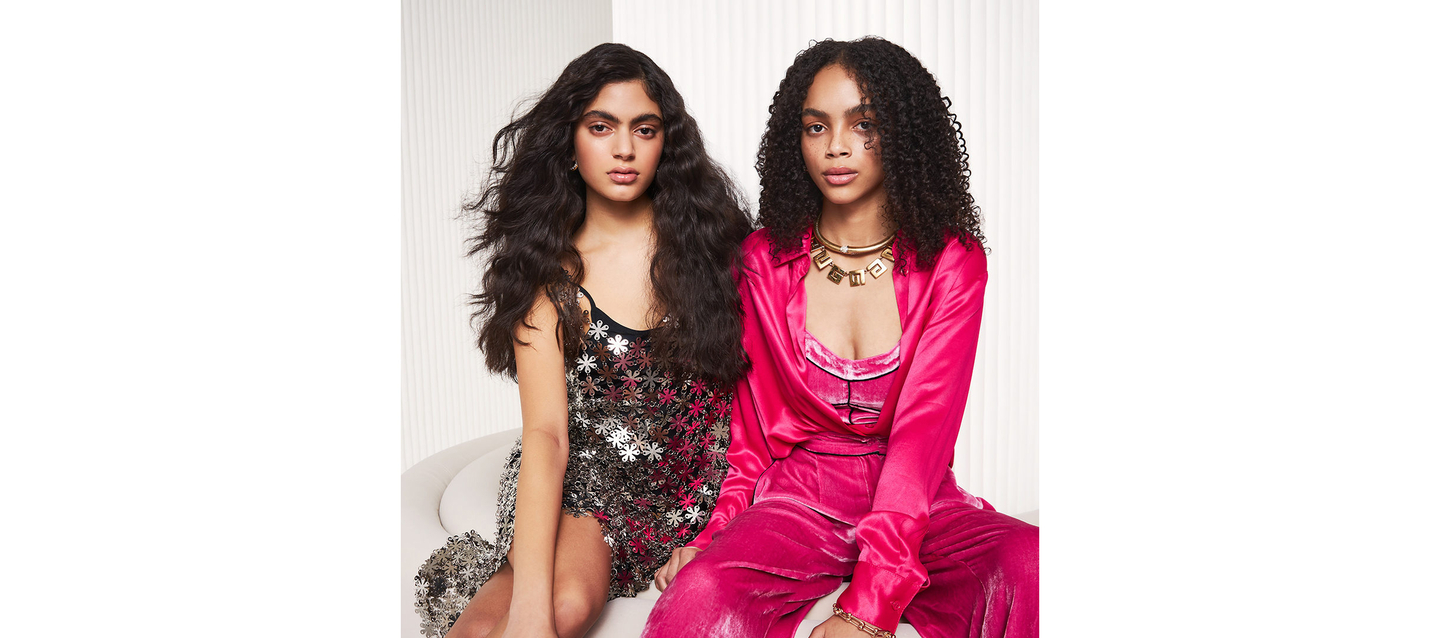 Two women with different curly hairstyles sitting together, one in a sequined dress and the other in a pink satin suit.